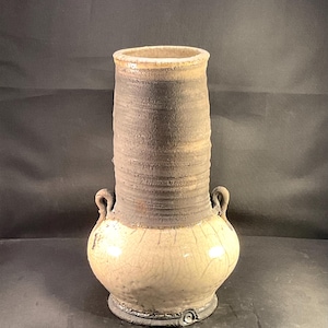 May include: A tall ceramic vase with a two-tone finish. The upper cylindrical neck features a dark, textured, matte gray surface with horizontal ridges, while the rounded lower body has a smooth, cream-colored glaze with a fine crackle pattern.