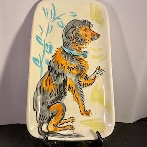 Puede incluir: Bandeja rectangular de cerámica con una ilustración de perro pintada a mano. El perro está representado en naranja, gris y negro, con un collar azul. El fondo es blanquecino con detalles azules y amarillos. Soporte de metal negro.