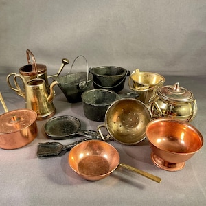 Puede incluir: Una colección de miniaturas de utensilios de cocina, que incluye ollas y sartenes de cobre, una tetera de latón y pequeños cubos de metal. Los artículos varían en color, desde cobre hasta latón y gris oscuro. La disposición está sobre una superficie gris.