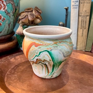 Peut inclure: Un pot en céramique avec une finition marbrée aux motifs tourbillonnants orange, verts et blancs. Le pot présente un corps arrondi, une large ouverture avec un bord lisse et un intérieur beige clair. La surface extérieure affiche un design fluide.