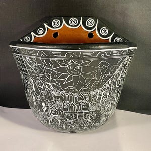 Peut inclure: Poche murale en céramique noire avec une illustration blanche détaillée d'une scène de village, dont un soleil souriant. Le bord supérieur présente un motif décoratif avec deux trous. L'intérieur est marron.
