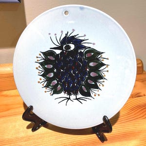 Könnte beinhalten: Dekorative weiße Keramikplatte mit handgemaltem Pfauenmuster. Der Pfau hat blaue, grüne und violette Details. Die Platte hat ein Loch zum Aufhängen und wird auf einem schwarzen Metallständer ausgestellt.