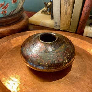 May include: A low, rounded ceramic vase with a dark, mottled glaze featuring shades of brown, green, and metallic copper. The vessel has a smooth, slightly uneven surface texture and a small, circular opening at the top, centred on a flat base.