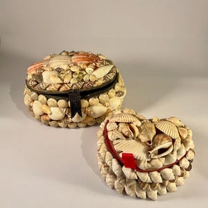 May include: Two decorative boxes covered in seashells. One is round with a hinged lid and a black ribbon closure. The other is heart-shaped with a red ribbon accent. The boxes are adorned with various sizes and colours of seashells.