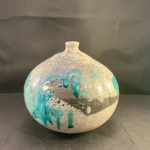 May include: A round ceramic vase with a narrow neck, featuring a crackled off-white glaze finish. The surface is decorated with abstract splashes of vibrant turquoise blue and textured matte black accents that create a striking contrast.
