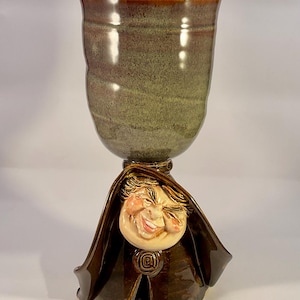 May include: A handmade ceramic goblet with a whimsical design. The goblet features a brown and green glazed finish, with a smiling face sculpted at the base. The goblet is approximately 7 inches tall.