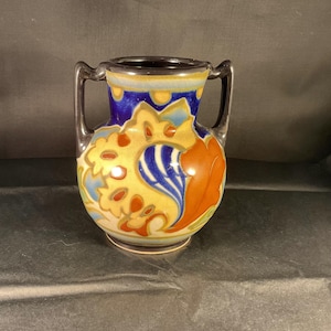 Puede incluir: Un pequeño jarrón decorativo con dos asas. El jarrón presenta un colorido diseño floral con detalles en amarillo, azul, naranja y rojo. El borde y las asas son de color gris oscuro. El jarrón es probablemente de cerámica o porcelana.