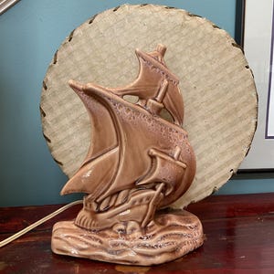 Może przedstawiać: Ceramiczna lampa stołowa z błyszczącą brązową rzeźbą statku na tle okrągłego, teksturowanego beżowego klosza z obszytą krawędzią. Podstawa przedstawia stylizowane fale w pasującym brązowym szkliwie, z białym przewodem elektrycznym z boku.