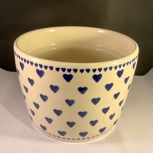 Puede incluir: Maceta de cerámica color crema con un patrón repetido de corazones azules. El borde presenta una fila de pequeños corazones azules. La maceta tiene una forma ligeramente acampanada y un acabado liso y brillante.