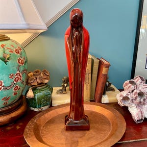 May include: A tall, slender ceramic figurine featuring a stylized figure with hands clasped in a prayer-like gesture. The sculpture has a glossy, multi-toned glaze transitioning from deep dark brown at the base to a vibrant, bright red at the top.