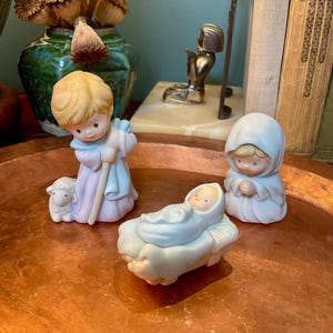 May include: A porcelain nativity figurine set featuring three figures: a standing shepherd in a lavender robe with a small white lamb, a kneeling figure in a light blue hooded cloak with hands clasped, and a baby wrapped in a light blue blanket.