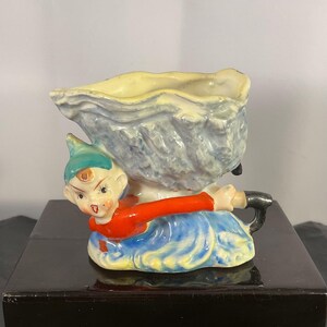 Könnte beinhalten: Vintage Keramikfigur eines sitzenden Elfen, der einen muschelförmigen Behälter hält. Der Elf trägt ein rotes Oberteil, einen blauen Rock und einen grünen Hut. Der Behälter ist hellblau und gelb.