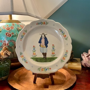 Könnte beinhalten: Ein weißer Keramikteller mit gewelltem Rand und einer handgemalten Illustration einer Person mit breitkrempigem Hut, blauer Jacke und heller Kniebundhose, flankiert von zwei farbenfrohen Blumenmotiven auf einem grünen Grasuntergrund.