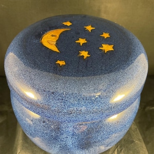 May include: A round, blue ceramic trinket box with a lid. The lid is decorated with a smiling yellow crescent moon and several yellow stars. The box has a speckled texture and a glossy finish, suggesting a handcrafted item.