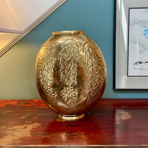 May include: A gold-coloured, oval-shaped vase with embossed grape and leaf designs. The vase has a wide base and a rounded top. The background includes a framed artwork and a red wooden surface.