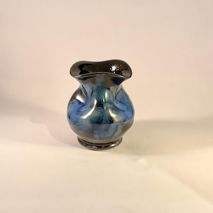 May include: A small ceramic vase with a distinctive form and a glossy finish. The vase has a dark, nearly black, rim that fades into shades of blue and teal. The base is a darker shade of blue.