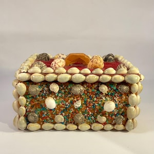 May include: A rectangular box decorated with seashells, small stones, and an orange-colored gem on the lid. The box is covered in a variety of seashells and small stones in shades of white, brown, and tan. The box is a decorative item.