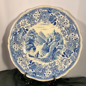 Puede incluir: Un plato decorativo azul y blanco con borde festoneado. El plato presenta un paisaje escénico con un castillo, un río y un borde floral. El plato está sostenido por un soporte de metal negro.