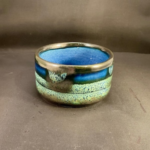 May include: A ceramic bowl with a vibrant color scheme. The interior is a deep blue, transitioning to a band of dark blue and green with a textured pattern. The rim is accented with a metallic silver.