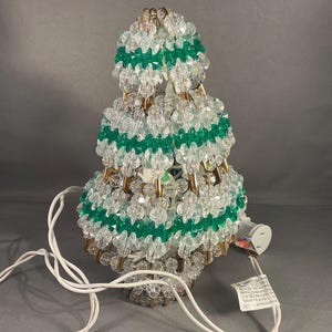 May include: A decorative Christmas tree-shaped lamp made of clear and green beads. The lamp has a white cord and plug. The tree is approximately 18 cm tall.