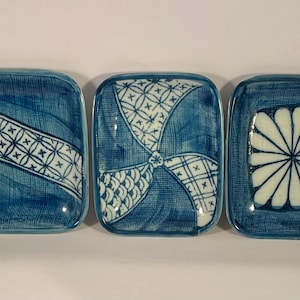 May include: Three rectangular ceramic dishes with blue and white patterns. Each dish has a unique design: a geometric band, a fan-like pattern, and a floral motif. These dishes are likely for serving or display.