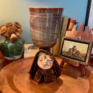 May include: A ceramic goblet featuring a tall, mottled brown and grey cylindrical cup resting on a base sculpted as a hooded figure with a smiling face. The figure wears a dark brown cloak with a circular emblem at the centre of the chest.