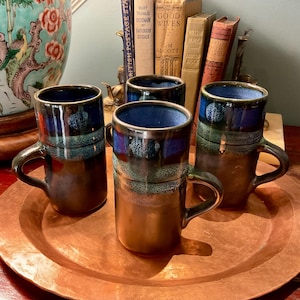 Può includere: Un set di tre tazze alte in ceramica con base marrone scuro e un motivo a smalto gocciolante blu e verde acqua vicino al bordo. Le tazze presentano una forma cilindrica con un manico robusto e sono disposte su un grande vassoio rotondo martellato.
