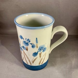 May include: White ceramic mug with a blue rim and base, decorated with blue iris flowers and brown stems. The mug has a curved handle and a smooth finish. A floral design for your kitchen, perfect for tea or coffee.