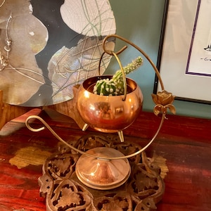 Peut inclure: Un petit pot en métal de couleur cuivre, rond et doté de trois pieds courts, suspendu à un support en métal doré courbé orné d'une rose décorative. Un cactus vert épineux est planté dans le pot, qui repose sur une base circulaire.