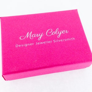 Puede incluir: Una caja de joyer&iacute;a rosa con el texto "Mary Colyer Designer Jeweller Silversmith" impreso en blanco.