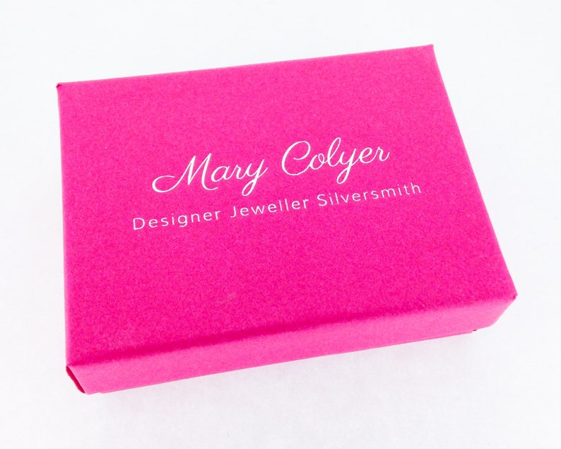 Puede incluir: Una caja de joyer&iacute;a rosa con el texto "Mary Colyer Designer Jeweller Silversmith" impreso en blanco.
