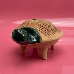 Puede incluir: Una figura de tortuga de cerámica con un caparazón marrón claro texturizado y una cabeza verde oscuro. La tortuga tiene tres patas pequeñas y un agujero en la parte superior. La figura está sobre un fondo rosa.