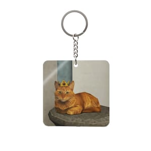 Puede incluir: Llavero cuadrado con un gato atigrado naranja que lleva una corona dorada con joyas rojas y verdes. El gato está tumbado sobre una superficie de piedra. El llavero tiene una cadena y un anillo plateados.