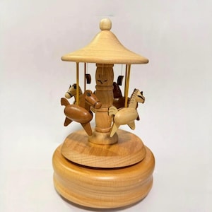Puede incluir: Caja de música de carrusel de madera con acabado marrón claro. El carrusel tiene un techo abovedado y cuatro caballos de madera. La base y la columna central también son de madera.