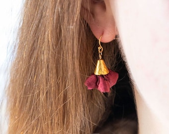 Puede incluir: Un par de pendientes de oro con un borla de tela color burdeos. Los pendientes cuelgan del lóbulo de la oreja.