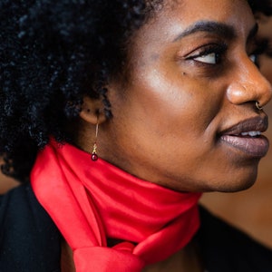 Op de afbeelding: Een vrouw met donker krullend haar draagt een rode sjaal om haar nek. Ze draagt een gouden oorring met een rode edelsteen.