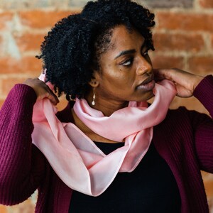 Op de afbeelding: Een persoon draagt een bordeauxrode cardigan met een roze zijden sjaal om de nek. De sjaal is in een losse knoop geknoopt en de uiteinden hangen naar beneden. De persoon heeft donker krullend haar en kijkt naar de zijkant.