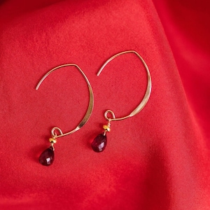 May include: A pair of gold-coloured hoop earrings with a teardrop-shaped, dark red gemstone pendant. The earrings are displayed on a red fabric background. The earrings have a simple, elegant design.