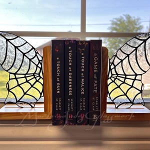 Könnte beinhalten: Buchstützen in Form von Schmetterlingsflügeln aus Glasmalerei und Holz. Die Flügel sind transparent mit schwarzen Konturen und gelben und weißen Akzenten. Die Buchstützen halten vier Bücher aus der Reihe "A Touch of Darkness".