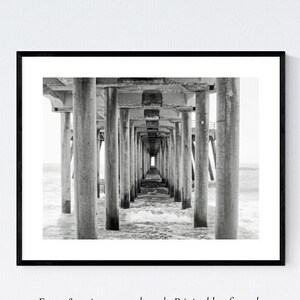 Pode incluir: Fotografia a preto e branco de um cais com vigas e pilares de madeira que se estendem sobre a &aacute;gua. A perspetiva &eacute; de baixo do cais, olhando para o fim.