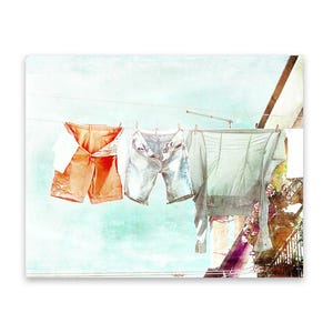 May include: A watercolor painting of clothing hanging on a clothesline. The clothing includes a pair of orange shorts, a pair of blue denim shorts, and a light gray shirt.