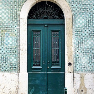 May include: A teal green double door with a decorative iron grate window above. The door is set in a white stone archway and surrounded by blue and white patterned tiles.