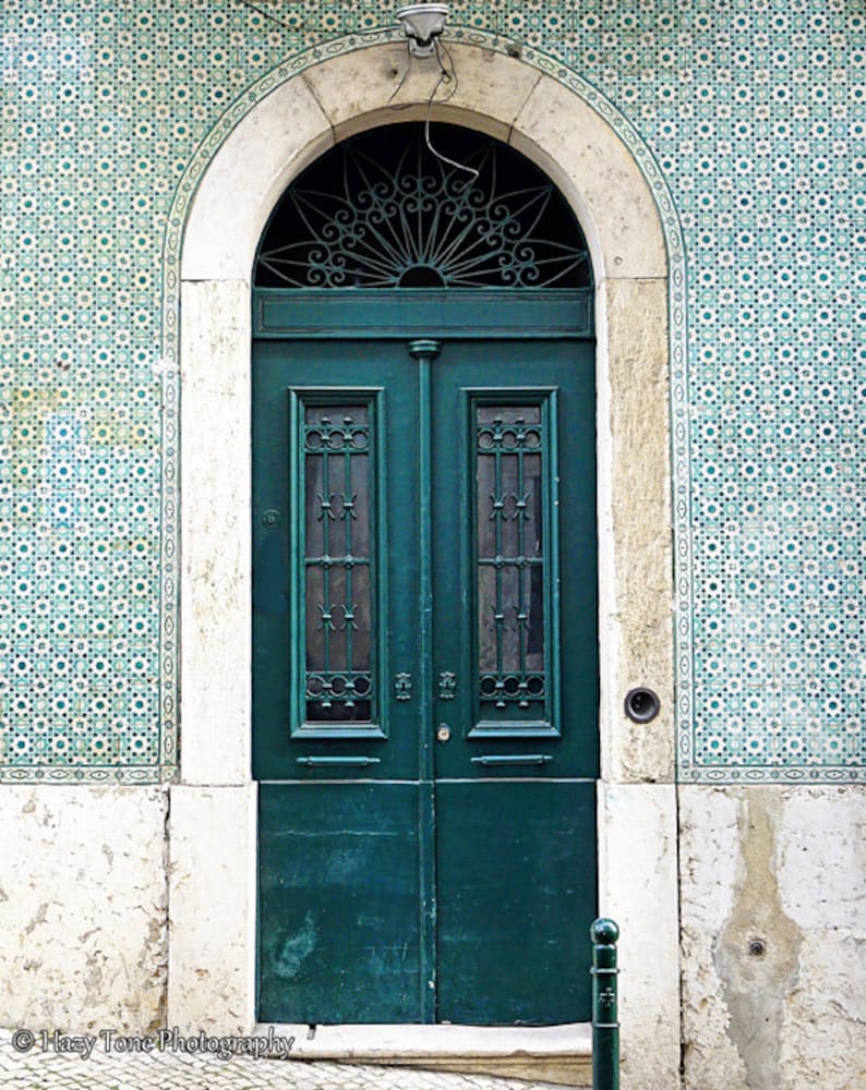 May include: A teal green double door with a decorative iron grate window above. The door is set in a white stone archway and surrounded by blue and white patterned tiles.