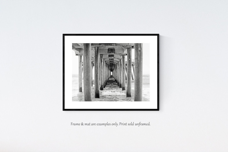 Pode incluir: Fotografia a preto e branco de um cais com vigas e pilares de madeira que se estendem sobre a &aacute;gua. A perspetiva &eacute; de baixo do cais, olhando para o fim.