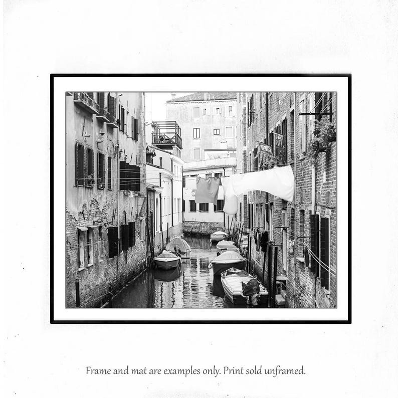 May include: A black and white photograph of a narrow canal in Venice, Italy. Buildings line the canal, and laundry hangs from lines strung across the water. Several small boats are moored in the canal.
