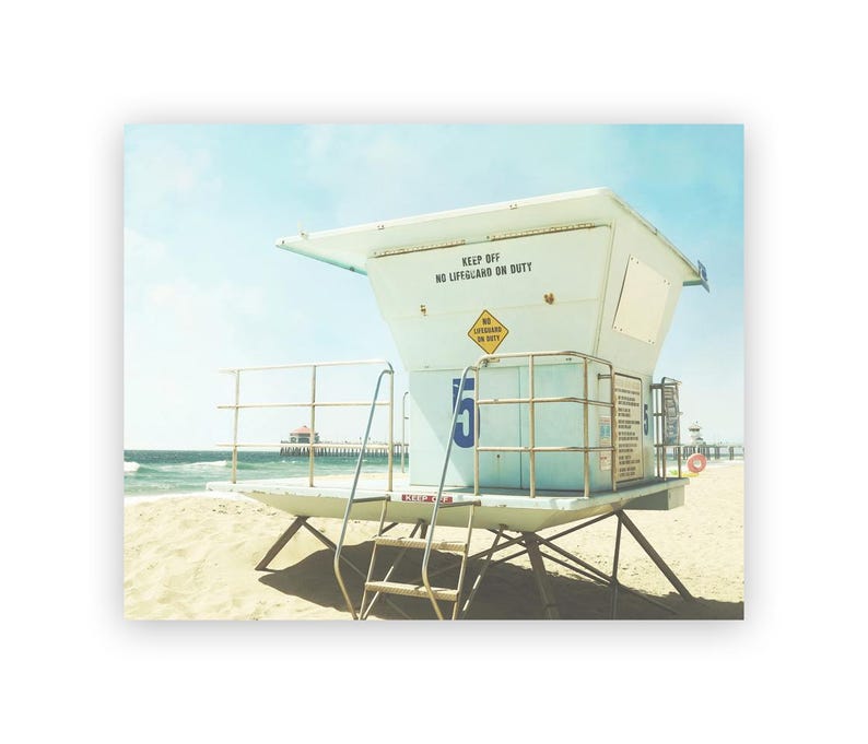 May include: A white lifeguard stand with a blue number "5" on the side. The stand has a sign that reads "Keep Off No Lifeguard On Duty". The stand is on a sandy beach with a pier in the background.