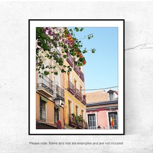 May include: A framed print featuring a vibrant street scene. Buildings in shades of peach, yellow, and red with wrought-iron balconies and window boxes. A Spanish flag hangs from a building, with a clear blue sky above.