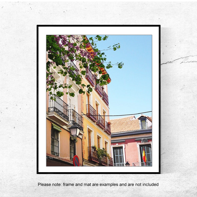 May include: A framed print featuring a vibrant street scene. Buildings in shades of peach, yellow, and red with wrought-iron balconies and window boxes. A Spanish flag hangs from a building, with a clear blue sky above.