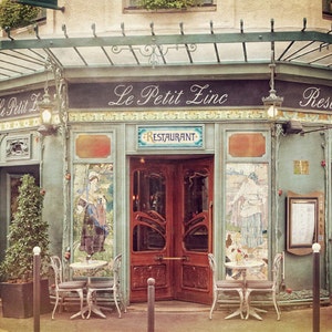 May include: A vintage Parisian cafe with a green awning and the words "Le Petit Zinc" and "Restaurant" on the facade. The cafe has two sets of doors, one with a glass panel and the other with a wooden panel. There are two tables with chairs outside the cafe.
