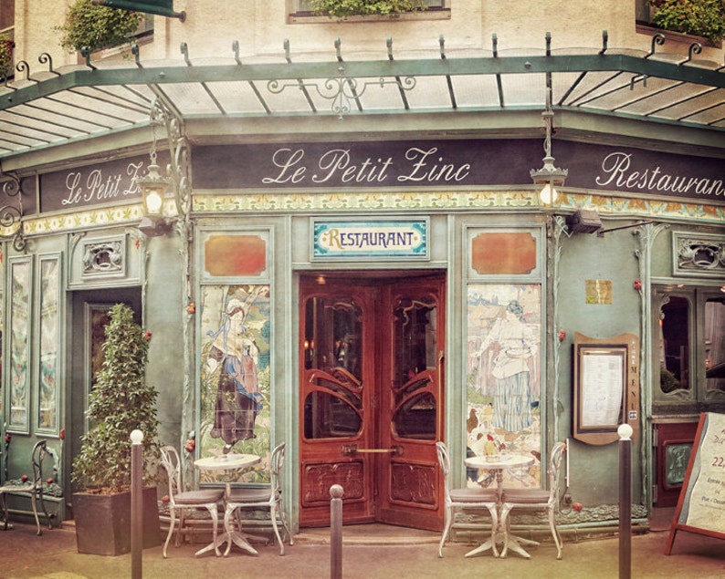 May include: A vintage Parisian cafe with a green awning and the words "Le Petit Zinc" and "Restaurant" on the facade. The cafe has two sets of doors, one with a glass panel and the other with a wooden panel. There are two tables with chairs outside the cafe.